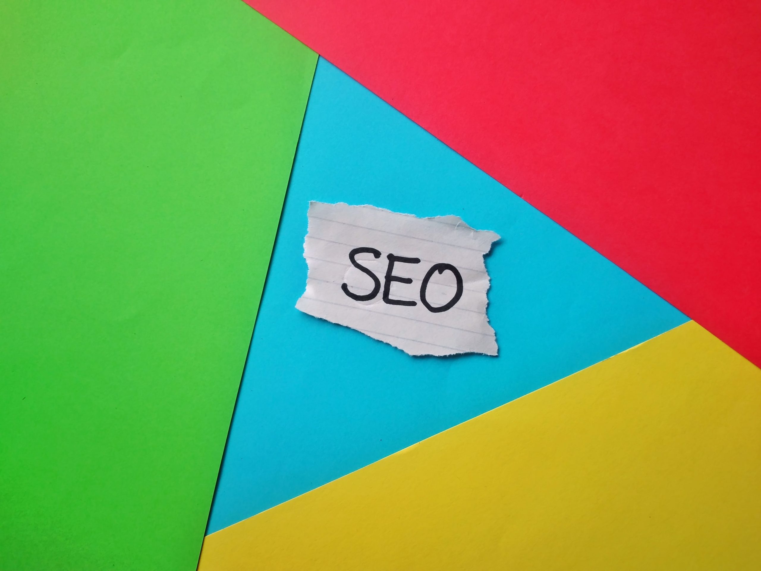 Imagen de un trozo de papel con la palabra SEO escrita, colocado sobre un fondo colorido de tonos verde, azul, rojo y amarillo, representando el concepto de optimización de contenidos para SEO.