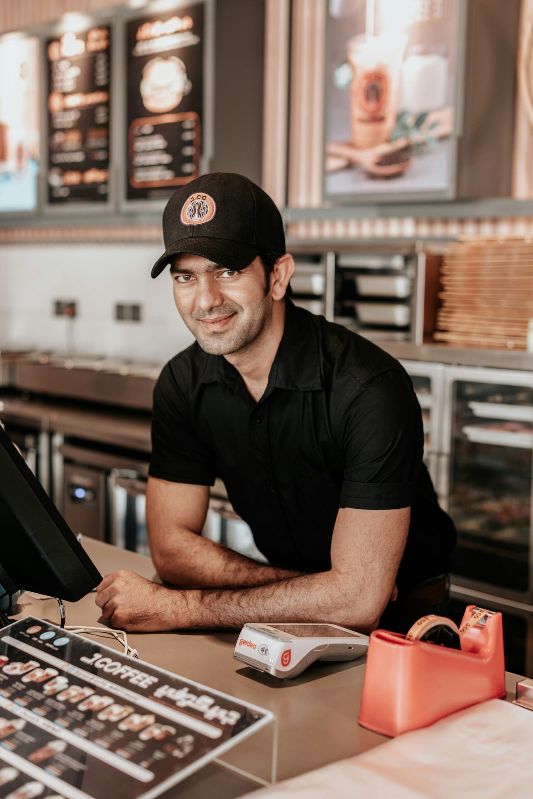 Empleado de cafetería con gorra y camisa negra apoyado en el mostrador con menús y datáfono, sonriendo a la cámara.