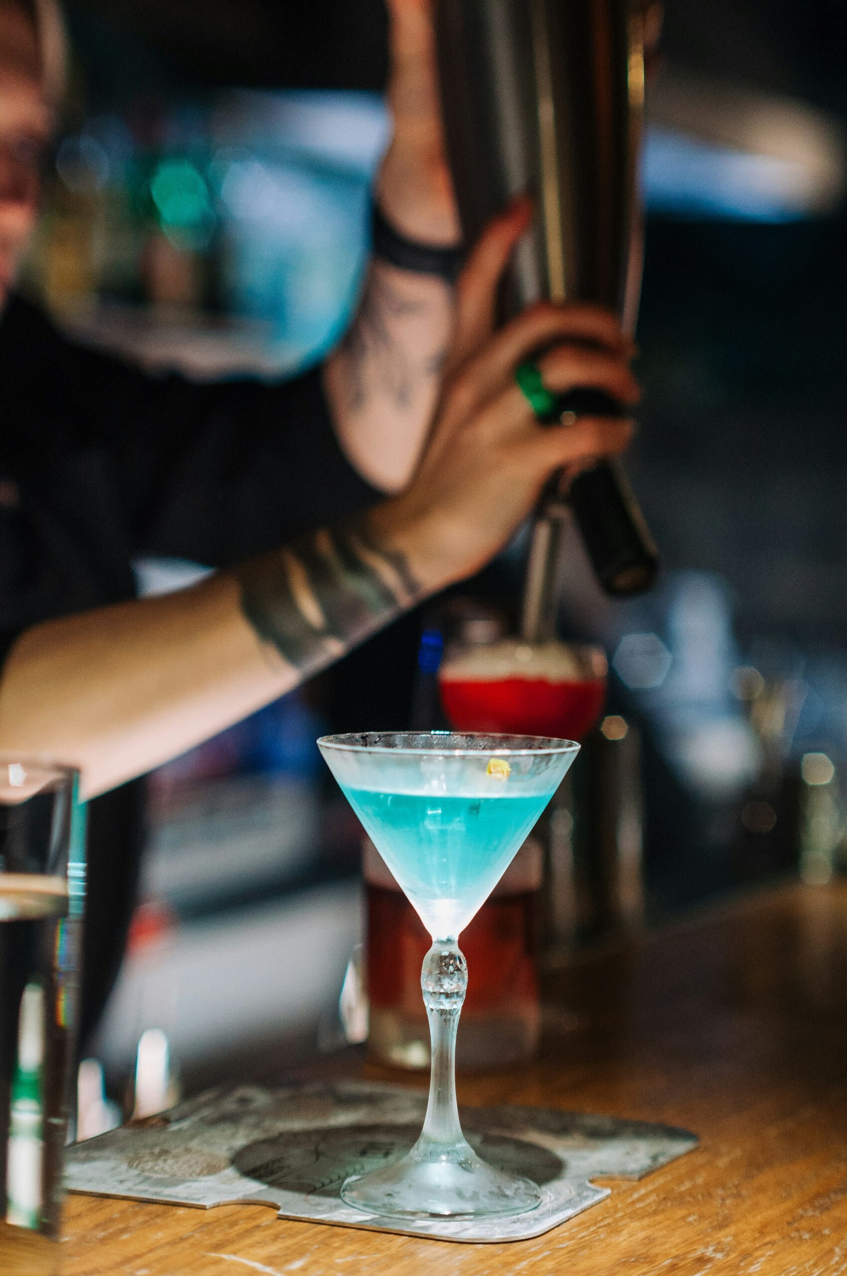 Copa de martini con bebida azul sobre posavasos en barra de madera, mientras bartender tatuado agita coctelera al fondo.