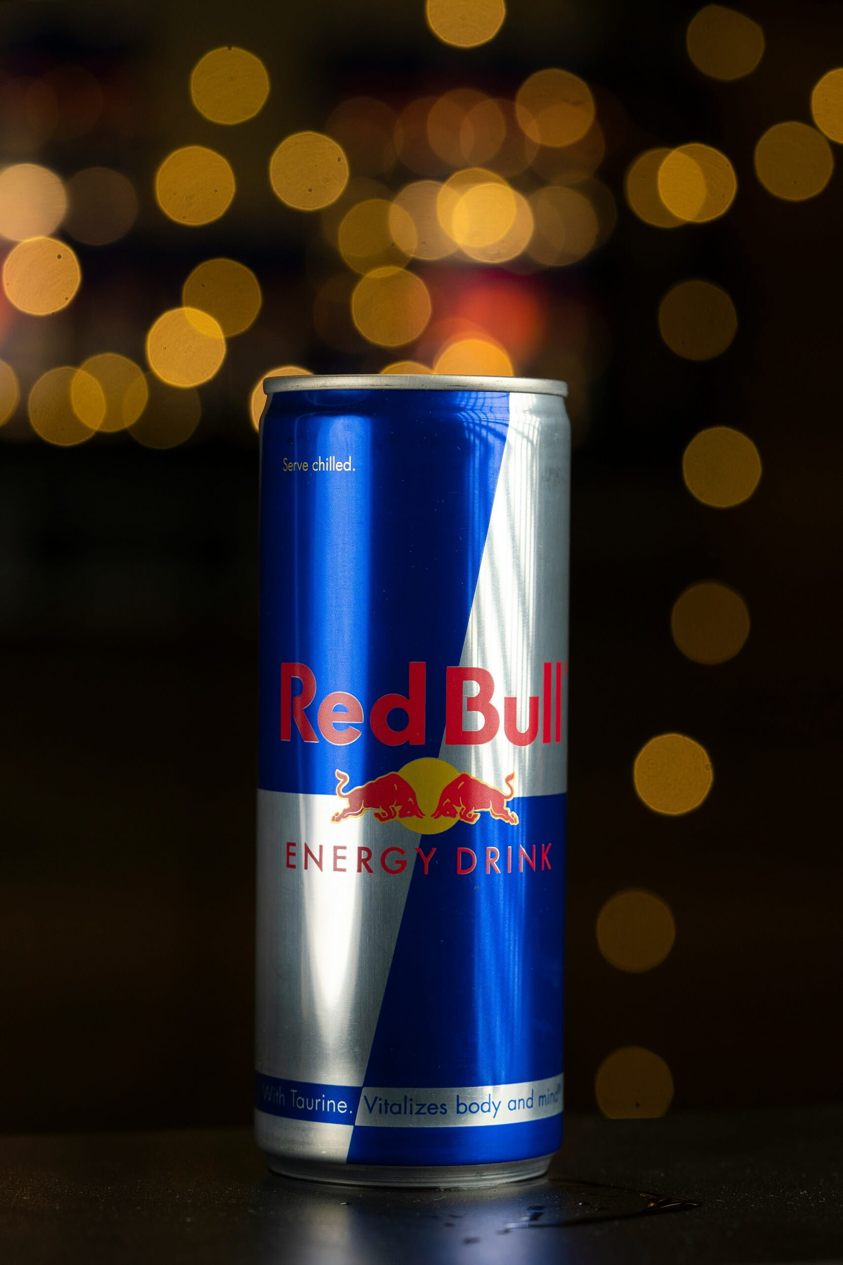 Lata metálica azul y plateada de Red Bull Energy Drink sobre una superficie oscura, con un fondo de luces cálidas desenfocadas.