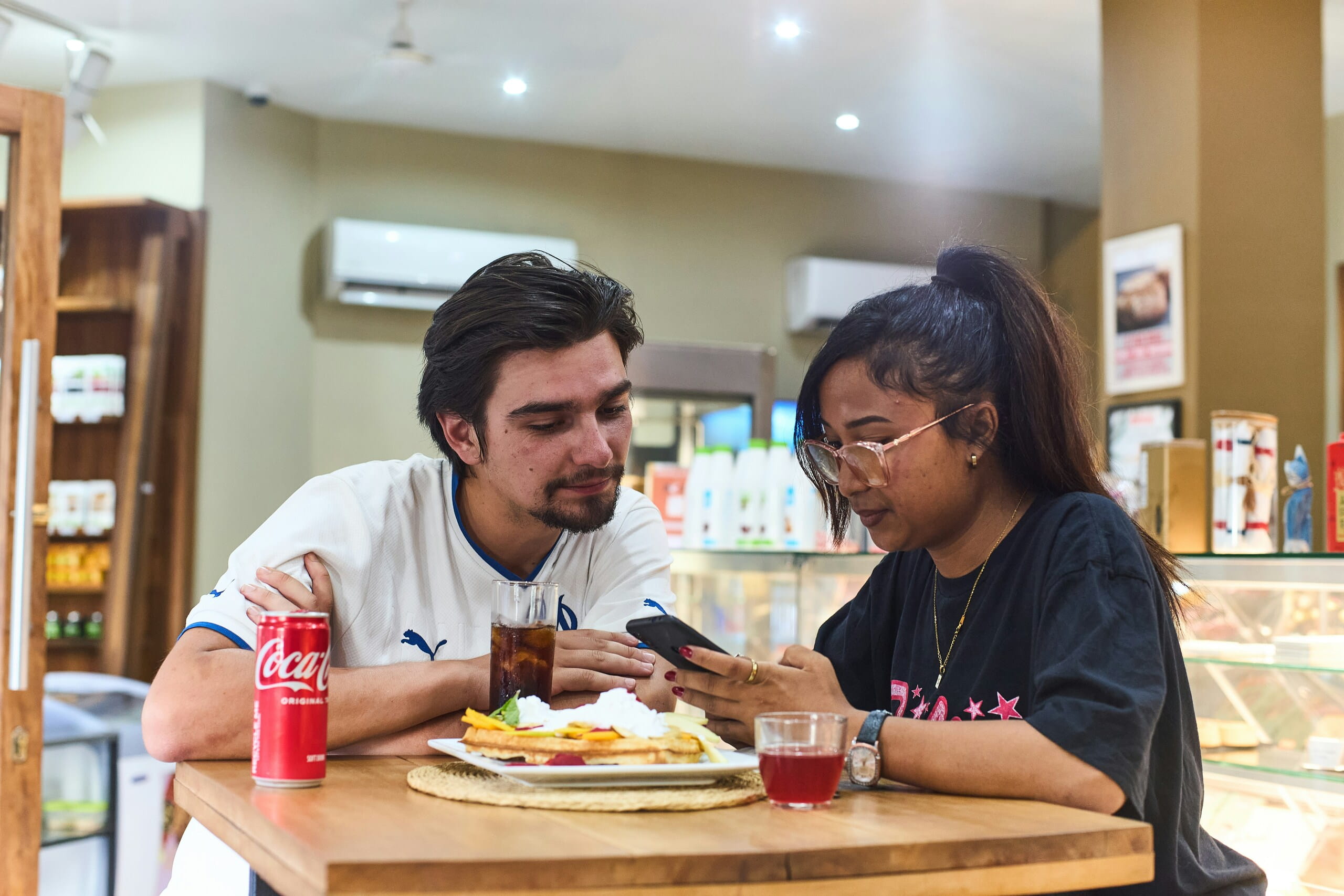 Un hombre y una mujer jóvenes miran juntos el teléfono móvil mientras desayunan en la barra de una cafetería.