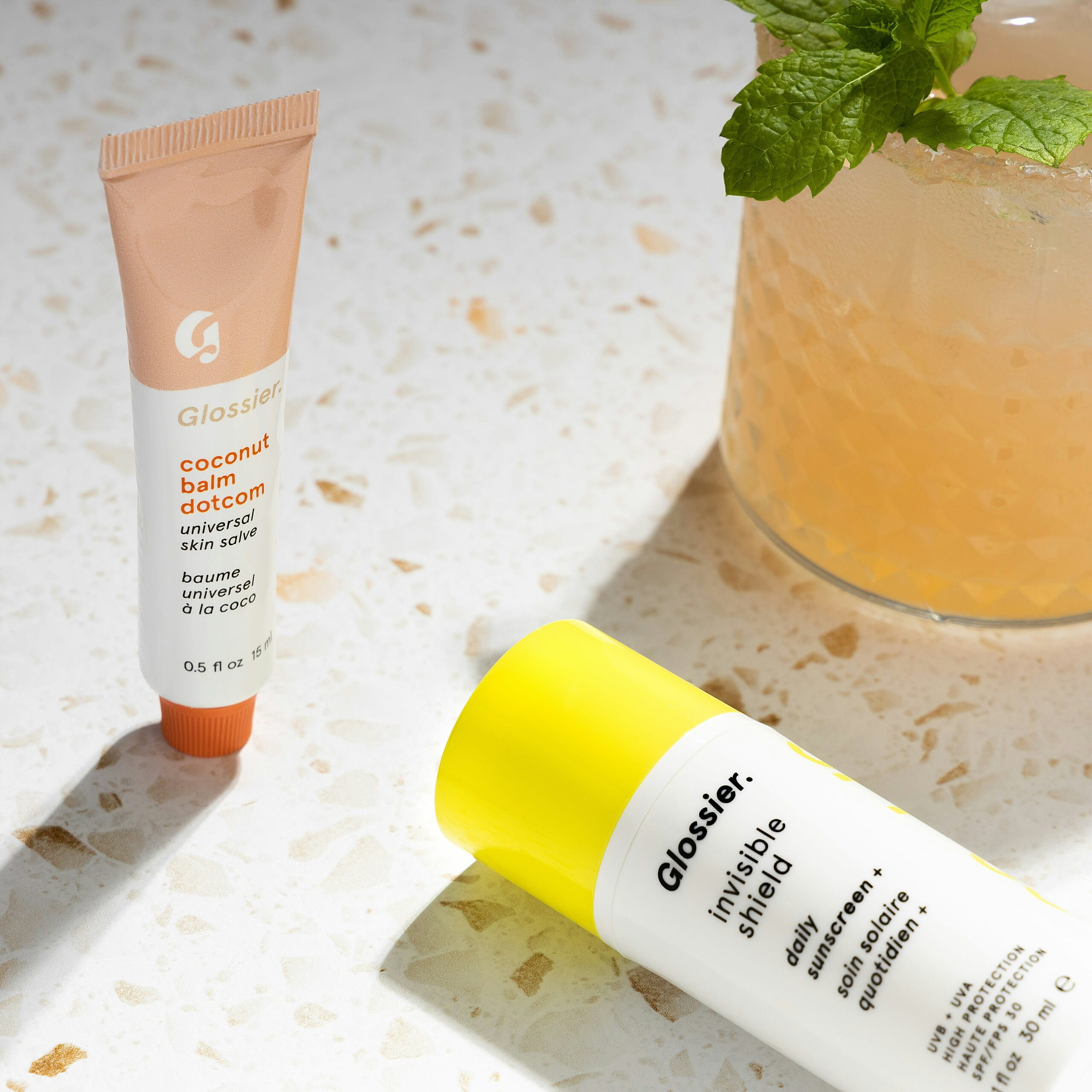 Tubo de bálsamo labial Glossier Coconut Balm Dotcom y envase de protector solar Invisible Shield junto a una bebida con menta.