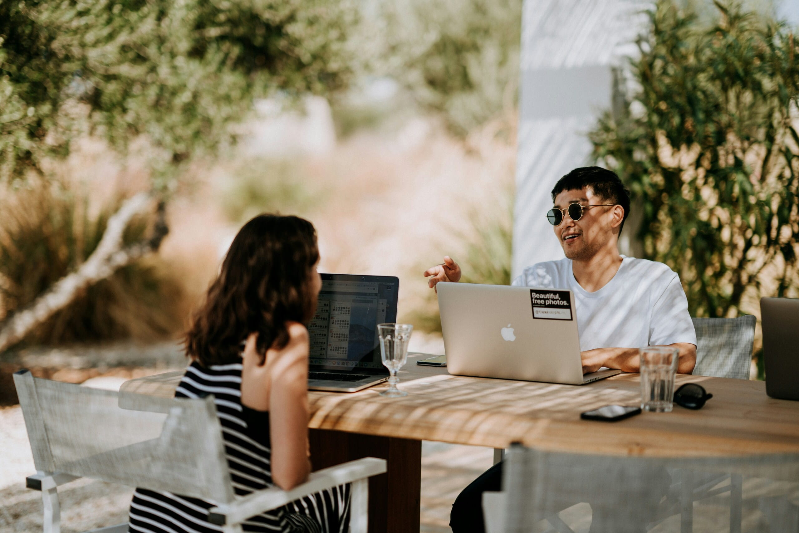 Trabajo remoto al aire libre con computadoras portátiles Dos profesionales trabajan en laptops en una mesa de madera bajo sombra de árboles en un espacio exterior.