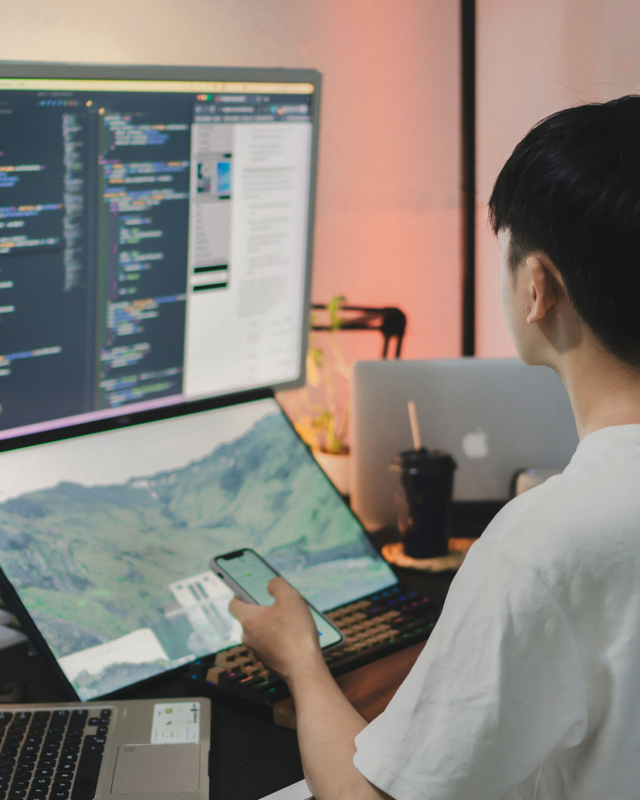 Joven programador usando un teléfono inteligente frente a un monitor con código y una pantalla portátil mostrando un paisaje.