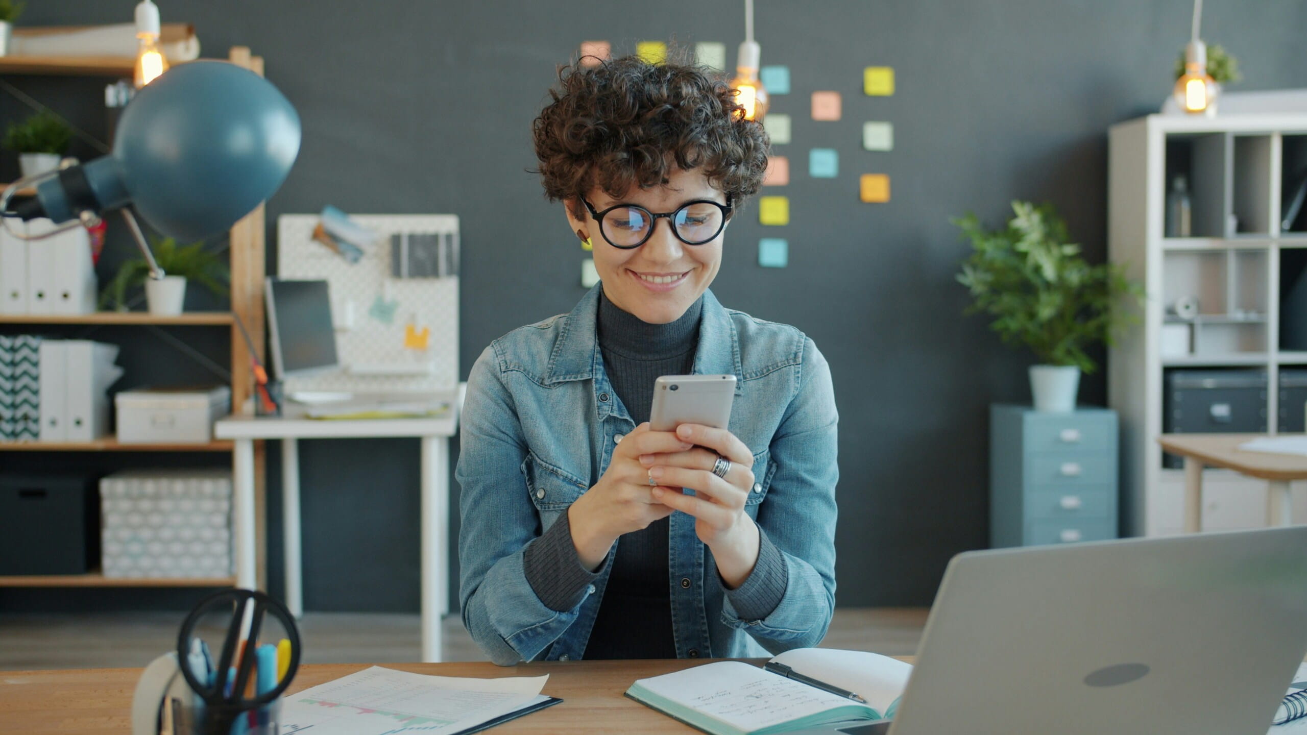 Mujer feliz con gafas y chaqueta vaquera usando su smartphone en escritorio con portátil y notas de colores.