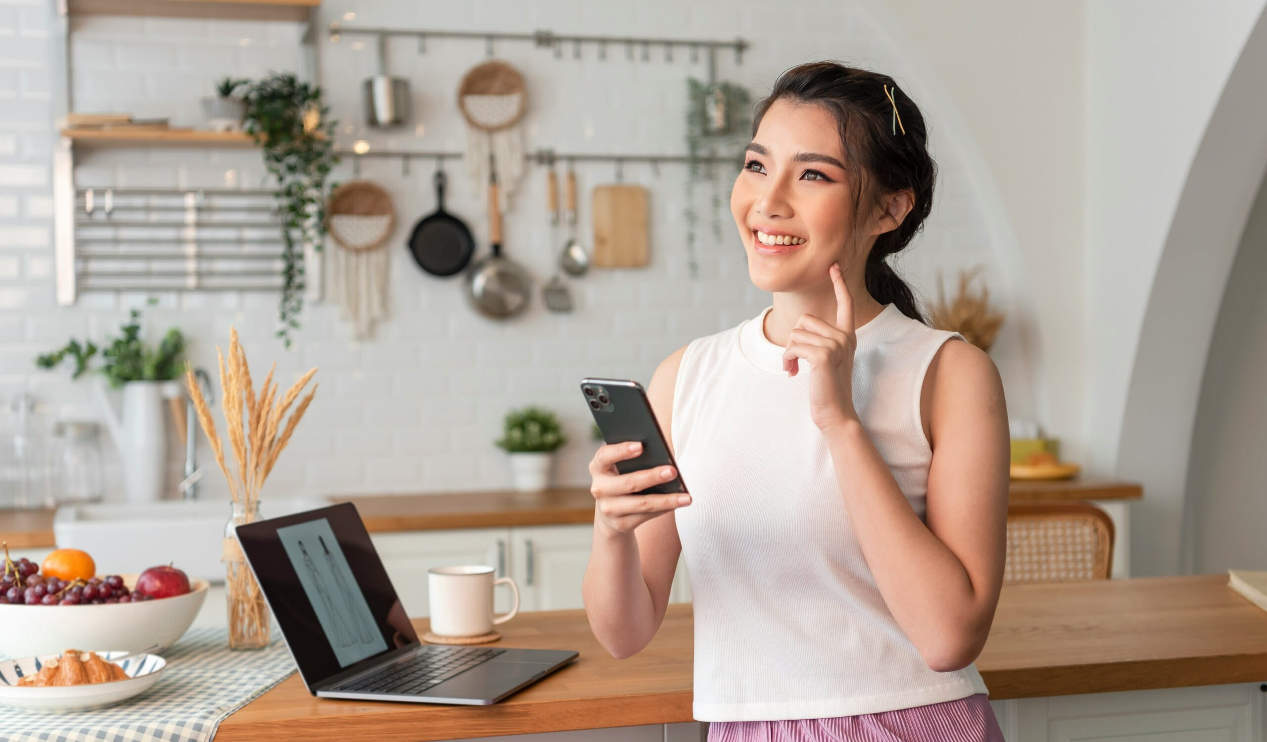 Mujer joven sonriente usando un smartphone en una cocina moderna con portátil y frutas.