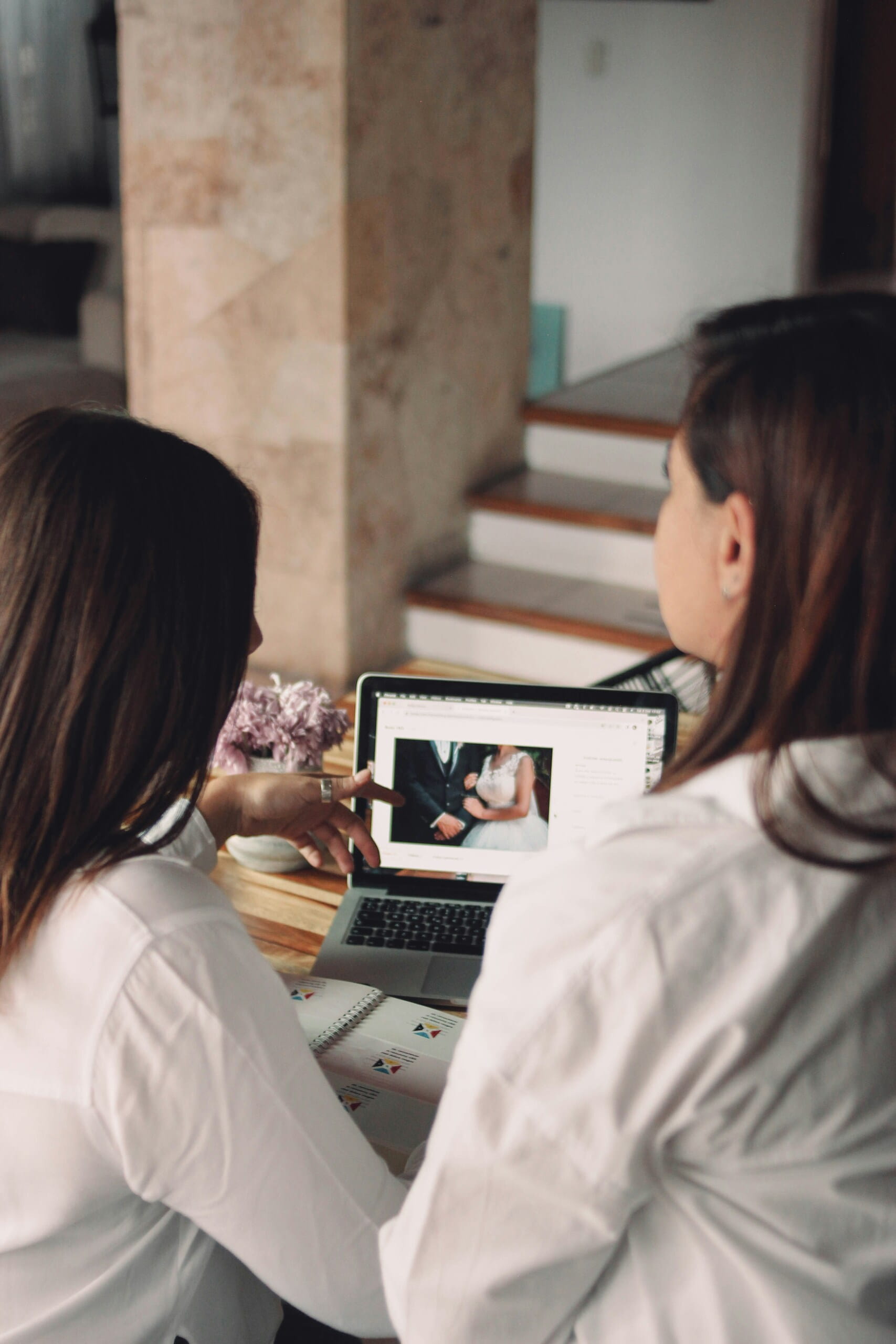 Dos mujeres señalando y revisando una fotografía de novios en la pantalla de una laptop durante una reunión.