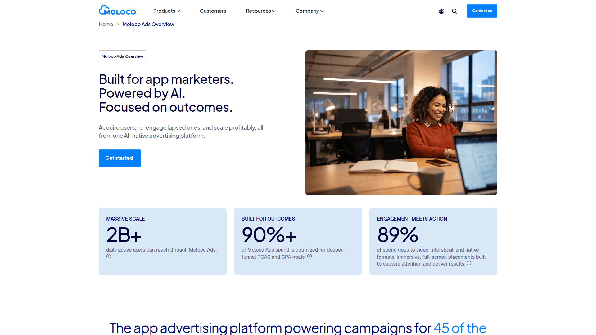 Mujer sonriente en una oficina moderna utilizando un ordenador portátil y un smartphone, que representa la plataforma de IA Moloco Ads para el marketing de aplicaciones.