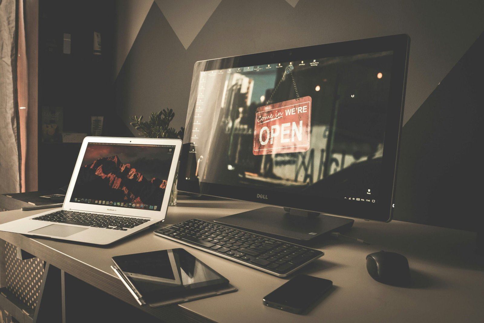 Puesto de trabajo de oficina en el que se ven un MacBook, un monitor Dell con el símbolo de «Abierto», un teclado negro, un ratón y dispositivos móviles.